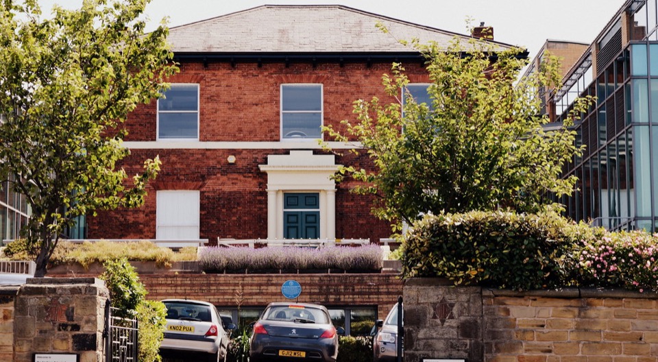 A red brick Victorian house behind a stone wall with cars parked outside